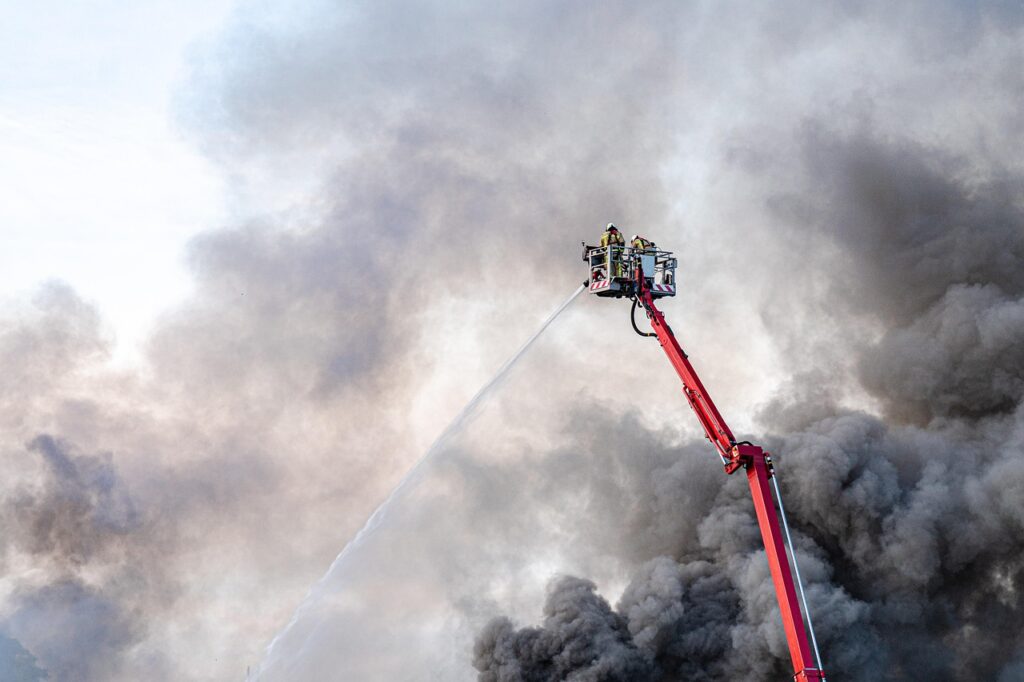 Bomberos en una grúa elevadora combaten un incendio entre densas nubes de humo.